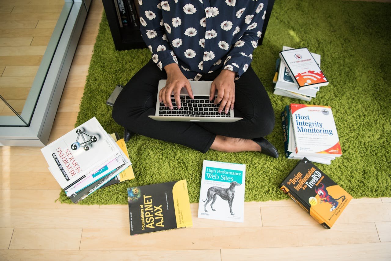 Woman sitting on green rug working on laptop, surrounded by technology books in a modern room.