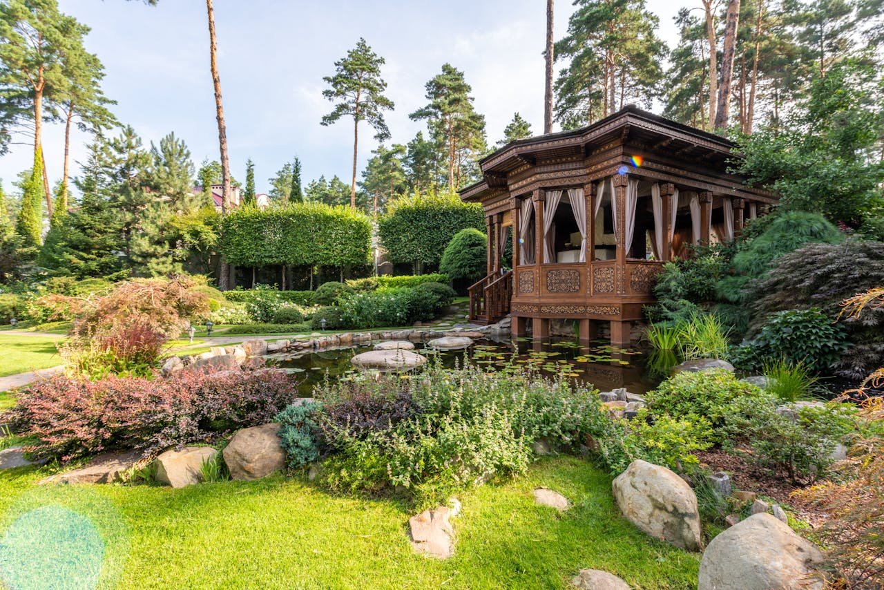 A peaceful wooden gazebo in a vibrant garden featuring a lush pond and diverse plants in summer.